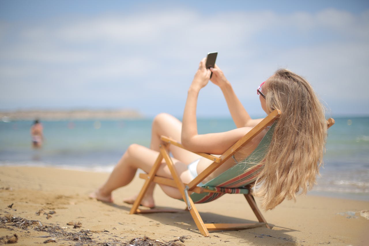 Blonde woman enjoying a sunny day on the beach, reclining in a chair with her smartphone.